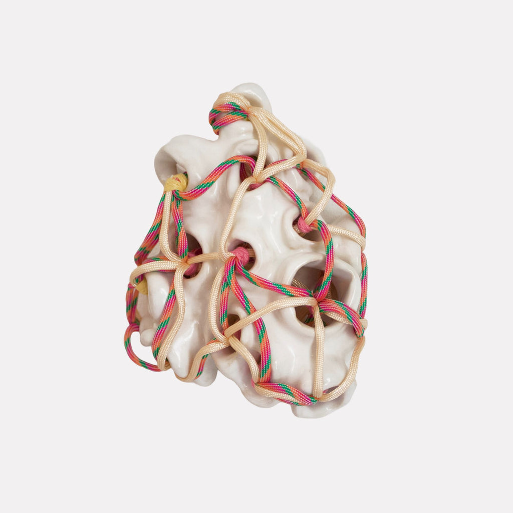 Within // Without by Hanna Washburn is a sculptural piece featuring a smooth, white organic form intricately intertwined with colorful woven cords in red, green, and beige tones. The tension between the rigid structure and flexible threads creates a compelling contrast, evoking themes of connection and containment.