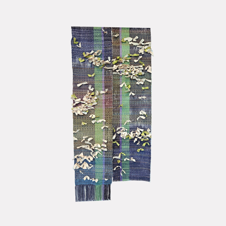 The artwork "Moth Cloth" by Johanna Ryan features a woven textile measuring 40 by 17.5 inches, combining hand-dyed fibers and textured Kitakata paper. The design showcases vertical stripes in deep blues, purples, and greens, accented by scattered curled paper pieces that add a dimensional, tactile quality. Johanna Ryan.