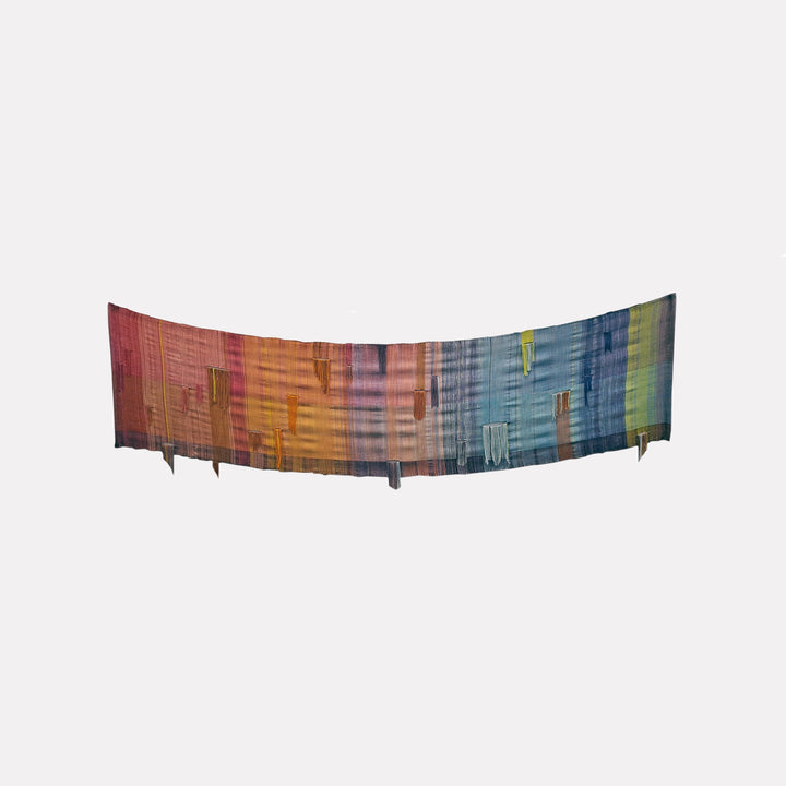The artwork "Rainbow Deconstruct" by Johanna Ryan features a wide, curved textile piece with a gradient of colors transitioning from red and orange to blue and green. It includes elongated vertical shapes and textures with an abstract, deconstructed style on fabric material. Johanna Ryan.