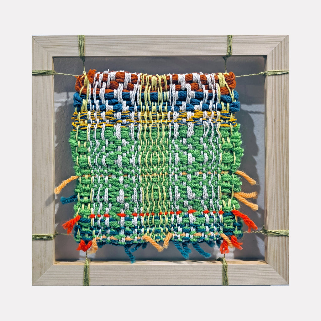The artwork "Interwork" features a vibrant woven textile with tightly interlaced yarns in green, blue, orange, white, and yellow, creating a textured, patterned square. It is framed in a simple wooden frame and showcases a handcrafted, artisanal style emphasizing colorful, woven materials. By Johanna Ryan.