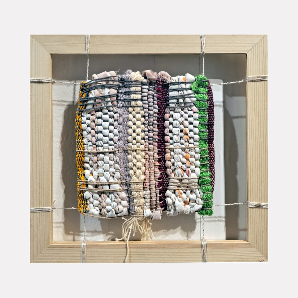 The artwork "Interworking" features a woven composition of cotton yarn in varied colors including white, beige, black, green, and yellow, arranged in vertical stripes with textured patterns and knots, framed in a light wooden square with intersecting strings. The piece measures 8 by 8 inches. By Johanna Ryan.