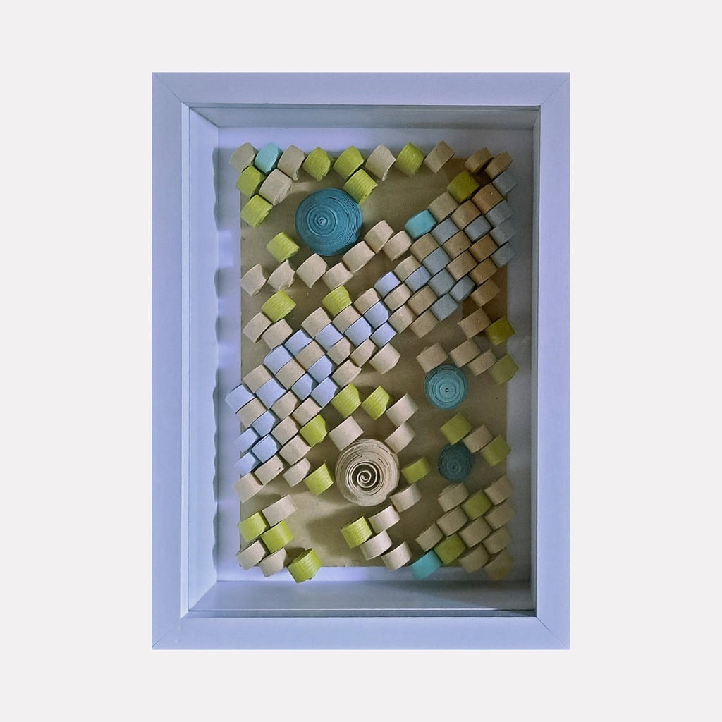 The artwork "Quilled" by Johanna Ryan features a framed 3D composition of small, rolled and folded paper pieces in shades of blue, green, cream, and beige, arranged in diagonal and spiral patterns that create a textured, geometric design with a delicate, handcrafted quilling style, highlighting intricate paper craft details.