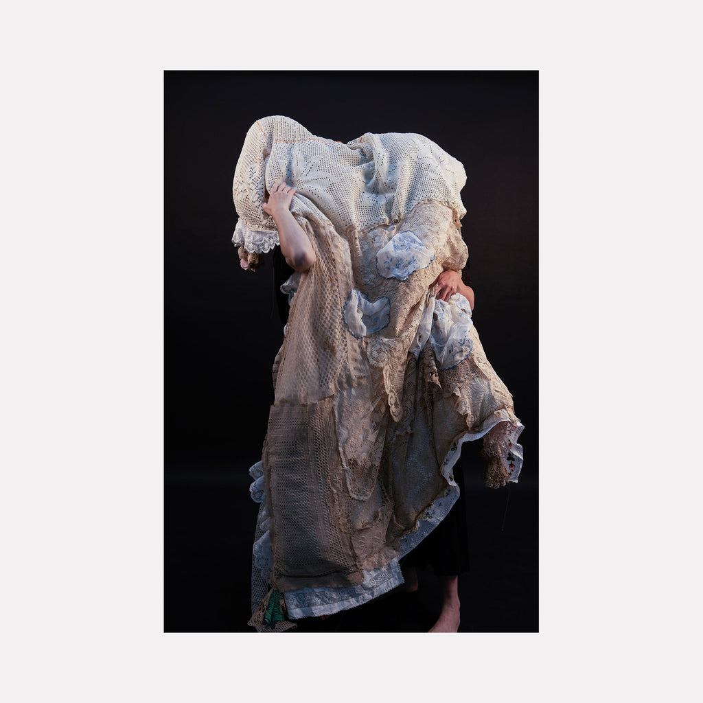 The artwork "080A8374" by Eunice Pais presents a figure enveloped in layered textiles of muted creams and off-whites, featuring varied textures like lace, mesh, and worn fabric. The sculptural form conveys a tactile intimacy and echoes the evocative textile work of Annette Messager and the ghostly figuration found in Kiki Smith’s pieces.