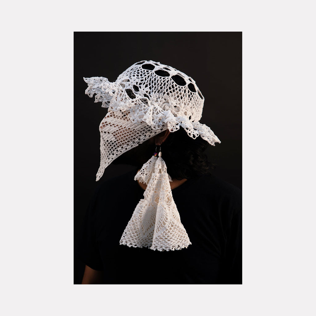 The artwork "080A8490" by Eunice Pais features a figure wearing an intricate white crocheted headpiece with open circular patterns and ruffled edges, complemented by delicate lace-like extensions hanging gracefully. The composition’s contrast against the black backdrop evokes the tactile artistry of Magdalena Abakanowicz and the intimate portraiture style of Cindy Sherman.