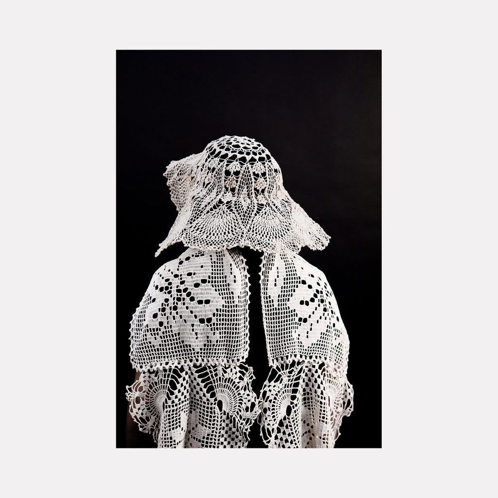 The artwork "080A8515" by Eunice Pais features a figure wearing an elaborate white crocheted ensemble with floral and geometric motifs, draping softly against a stark black backdrop. The intricate lacework and sculptural silhouette recall the textural refinement found in Louise Bourgeois’s fabric sculptures and the delicate craftsmanship of Eva Hesse’s fiber pieces.