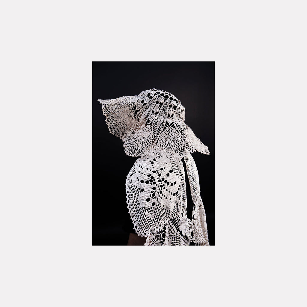 The artwork "080A8525" by Eunice Pais depicts a figure adorned in delicate white crocheted fabric featuring floral and lattice patterns, set against a solid black backdrop. The intricate, lace-like texture and sculptural form evoke the refined textile artistry reminiscent of Louise Bourgeois’s fabric works and the ethereal fiber explorations of Eva Hesse.
