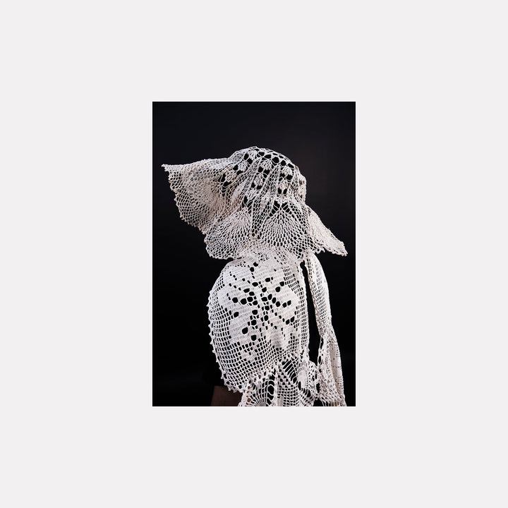 The artwork "080A8525" by Eunice Pais depicts a figure adorned in delicate white crocheted fabric featuring floral and lattice patterns, set against a solid black backdrop. The intricate, lace-like texture and sculptural form evoke the refined textile artistry reminiscent of Louise Bourgeois’s fabric works and the ethereal fiber explorations of Eva Hesse.