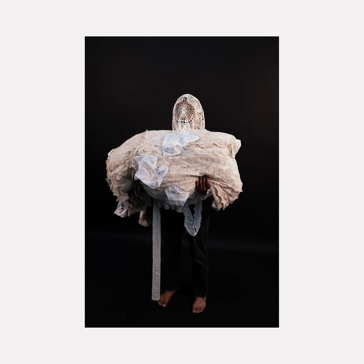 The artwork "080A8575" by Eunice Pais portrays a figure cloaked in layered, textured fabrics of soft whites and creams with intricate lace and mesh patterns, set against a stark black backdrop. The composition’s tactile quality and sculptural form evoke the intimate textile explorations of Annette Messager and the haunting presence reminiscent of Kiki Smith’s figurative pieces.