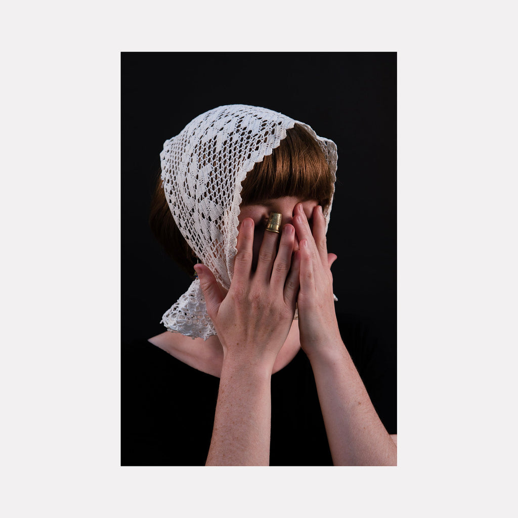 The artwork "080A8602" by Eunice Pais features a figure partially obscured by their hands, adorned with a white crocheted headscarf against a deep black background. The interplay of delicate textile patterns and pale skin tones evokes the intimate, tactile qualities found in the textile sculptures of Magdalena Abakanowicz and the quiet portraiture of Cindy Sherman.