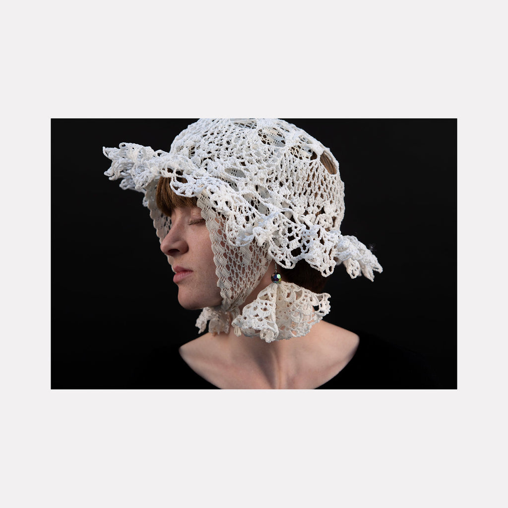 The artwork "080A8649" by Eunice Pais features a profile portrait of a figure wearing an intricately crocheted white bonnet with floral motifs and textured loops, set against a deep black background. The delicate craftsmanship and sculptural form recall the detailed textile work of Sheila Hicks and the ethereal quality of Eva Hesse’s fiber art.