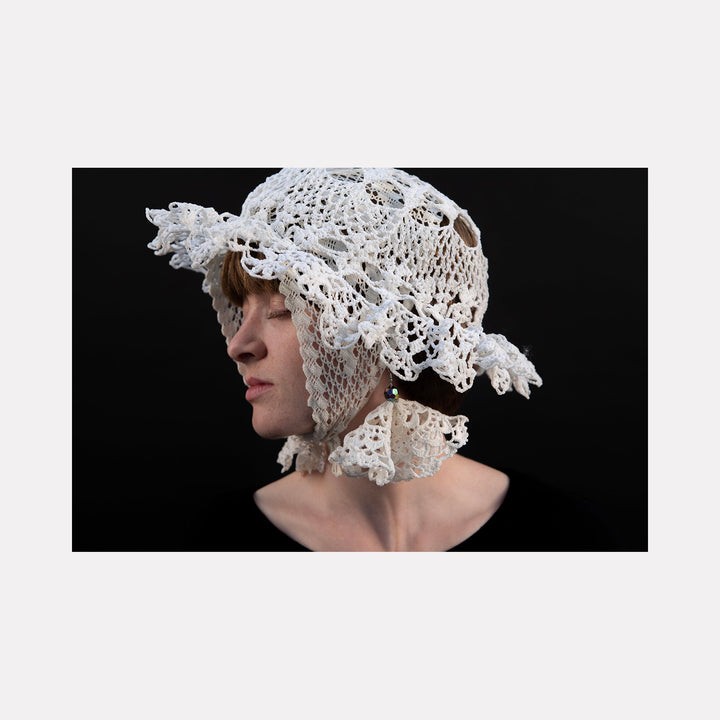 The artwork "080A8649" by Eunice Pais features a profile portrait of a figure wearing an intricately crocheted white bonnet with floral motifs and textured loops, set against a deep black background. The delicate craftsmanship and sculptural form recall the detailed textile work of Sheila Hicks and the ethereal quality of Eva Hesse’s fiber art.