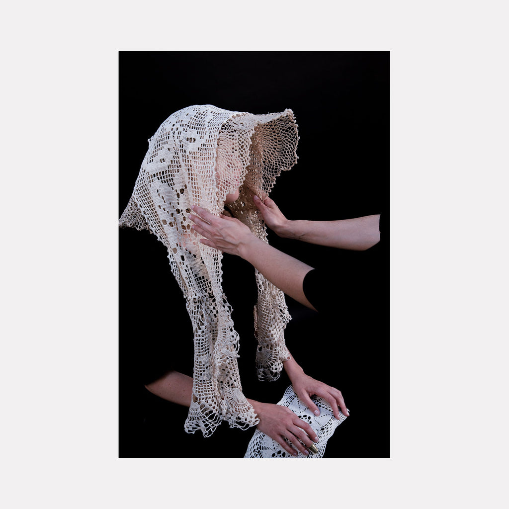 The artwork "080A8713" by Eunice Pais presents a delicate, intricately crocheted white lace garment draped over a figure’s head and shoulders. The soft texture and careful craftsmanship recall the tactile intimacy of Louise Bourgeois’s fabric works and the contemplative portraiture style of Cindy Sherman, set against a stark black background.