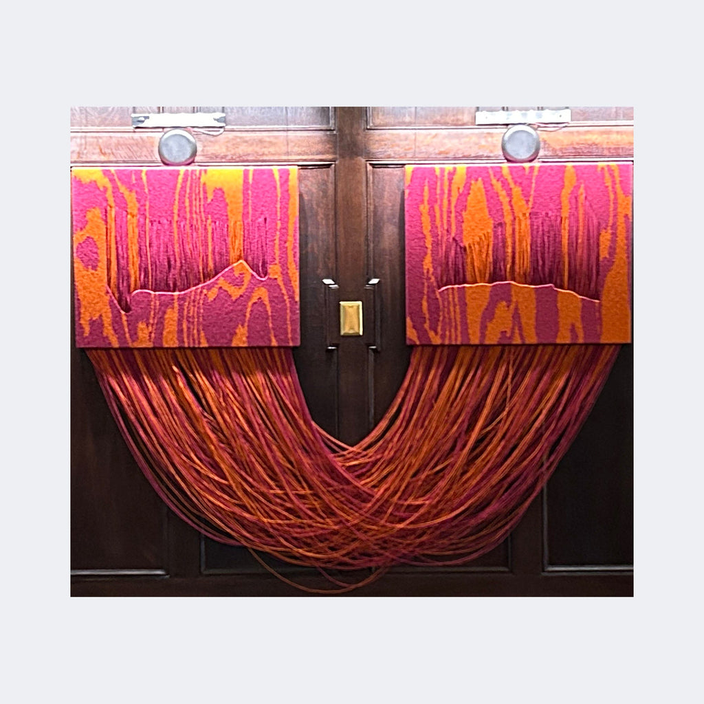 The artwork "Landlines II" by Quynn Douglas Dale features two rectangular panels with vibrant orange and pink abstract patterns resembling wood grain, mounted on a dark wooden background. Below, numerous flowing threads or strings in matching hues cascade and intertwine, creating a dynamic, tactile effect reminiscent of fiber art and echoing styles of artists like Sheila Hicks and Eva Hesse.
