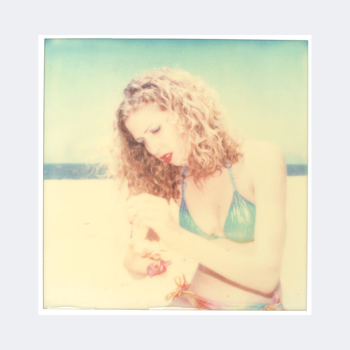 The artwork "Kelly (Beachshoot) 1374" by Stefanie Schneider depicts a soft-focused, vintage-style image of a woman with curly blonde hair wearing a teal bikini top on a sunlit beach. The pastel tones and faded textures evoke the dreamy, nostalgic feel of Polaroid photography, reminiscent of styles by artists like Sally Mann and Nan Goldin.
