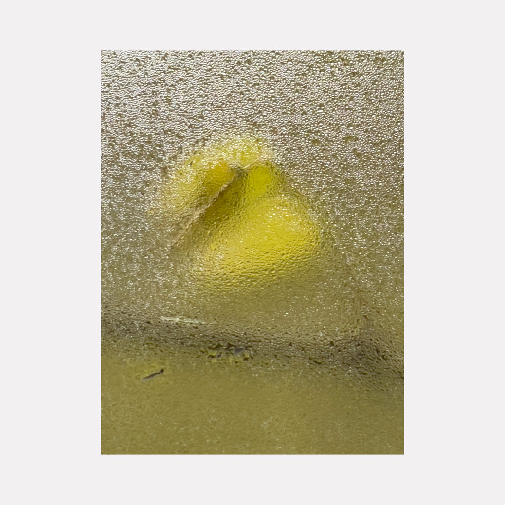 The artwork "Wisteria 2" by Molly Heron captures an abstract shape in muted yellow tones obscured by a textured, frosted surface. The composition emphasizes subtle light diffusion and grainy detail, evoking minimalist influences and the ethereal quality found in the works of Mark Rothko and Agnes Martin.