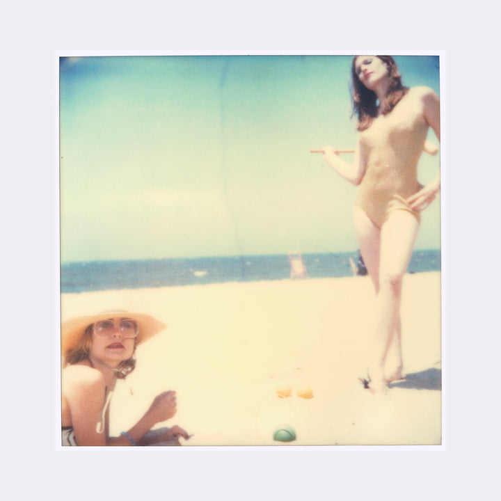 The artwork "Untitled (Beachshoot) 1410" by Stefanie Schneider features a soft-focused, vintage-style photograph with muted pastel tones capturing a beach scene. The composition includes two women, one reclining with a wide-brimmed hat and glasses, the other standing confidently in a swimsuit. The dreamy atmosphere and faded textures evoke the style of William Eggleston and Nan Goldin.