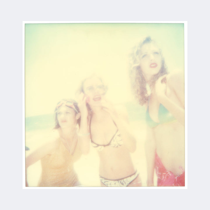 The artwork "Untitled (Beachshoot) 1418" by Stefanie Schneider depicts three women in swimwear standing by the beach, rendered in a soft-focus, dreamlike style with faded colors and nostalgic textures. The image evokes the vintage Polaroid aesthetic reminiscent of William Eggleston’s color photography and the hazy atmospheres found in some work by Nan Goldin.