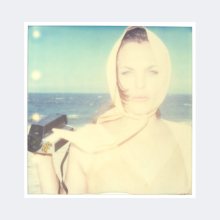 The artwork "Untitled (Beachshoot) 1461" by Stefanie Schneider features a softly blurred, vintage-style Polaroid photograph of a woman on a beach. She wears a pale scarf over her head, holding a retro camera, with muted pastel tones and light leaks reminiscent of William Eggleston’s color photography and the hazy dreaminess of early 20th-century photo portraits.