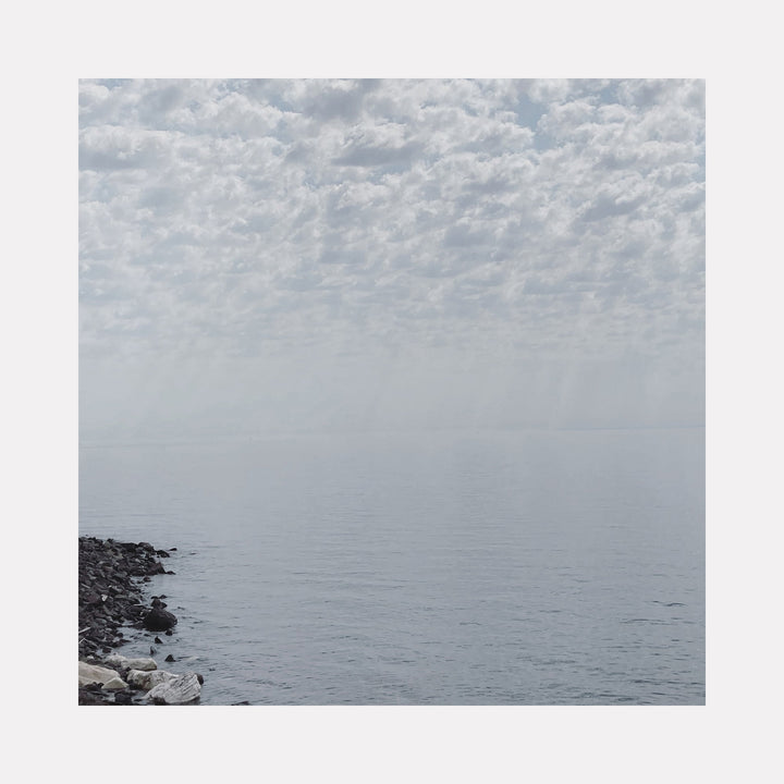 The artwork "White, Lake Superior" by Ann Tracy features a serene lakescape with soft, diffused light streaming through a textured, cloud-filled sky over calm water. The composition echoes the minimalist style of artists like Agnes Martin, emphasizing subtle shades of gray and delicate atmospheric effects, creating a tranquil, contemplative mood.