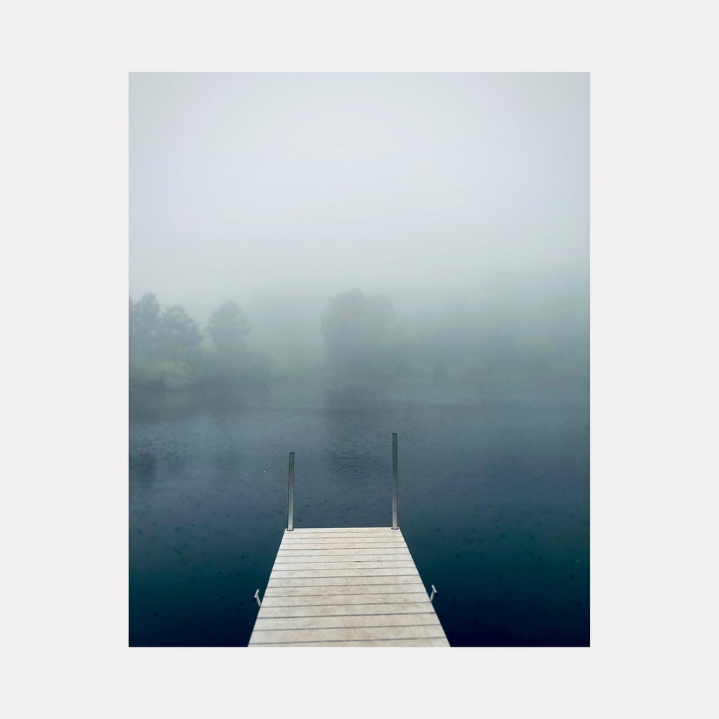 The artwork "Bagley Nature Area #1" by Ann Tracy depicts a serene white wooden dock extending into a dark, mist-covered lake with faint outlines of trees in the background. The soft, muted color palette and atmospheric fog evoke styles similar to Caspar David Friedrich’s romantic landscapes, emphasizing tranquility and introspection.