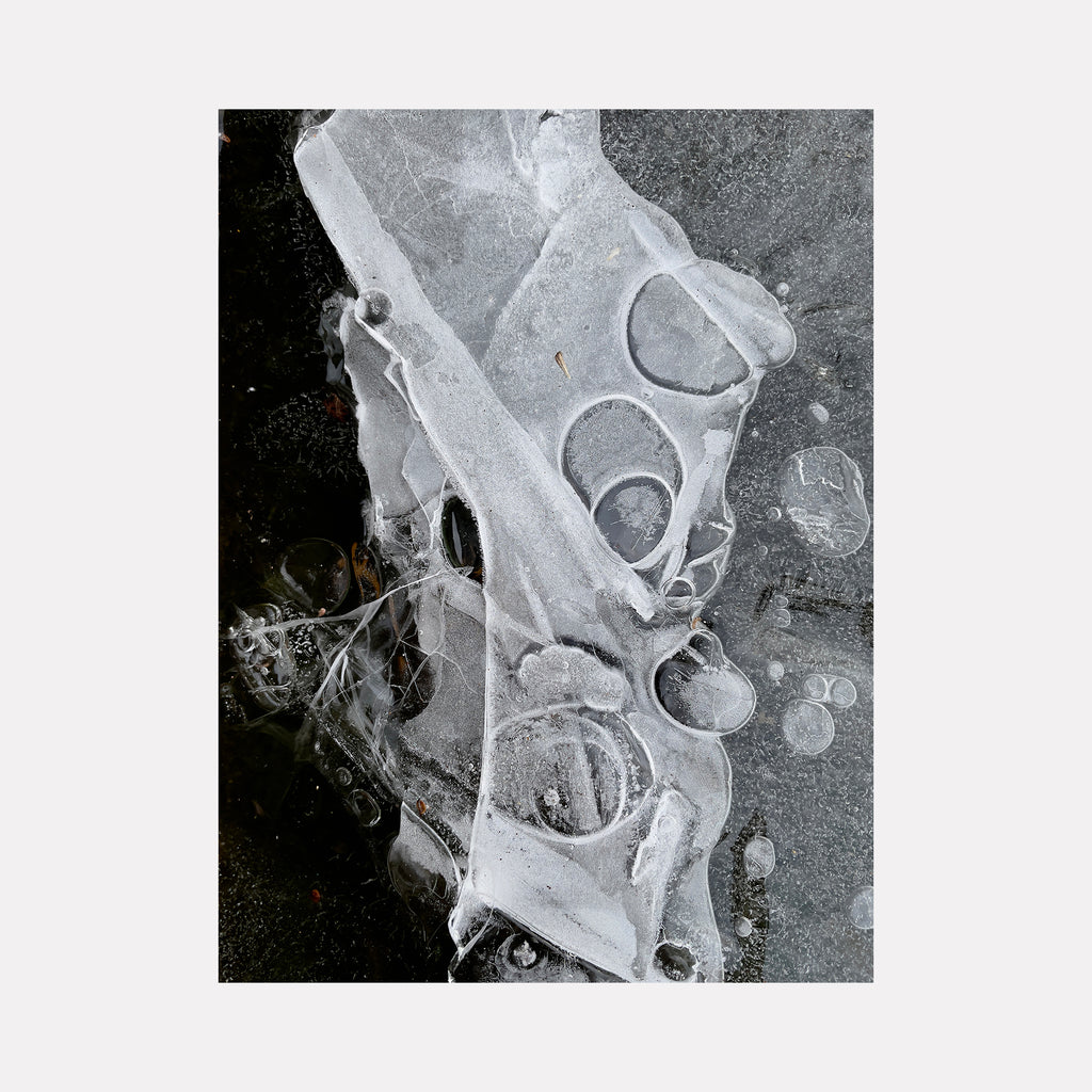 The artwork "Ice Bubbles" by Molly Heron captures a striking monochrome composition of frozen, translucent ice formations encasing circular air pockets and layered textures. The intricate interplay of light and shadow evokes the abstract fluidity found in Jasper Johns’ encaustic works and the natural precision reminiscent of Ansel Adams’ photographic studies.