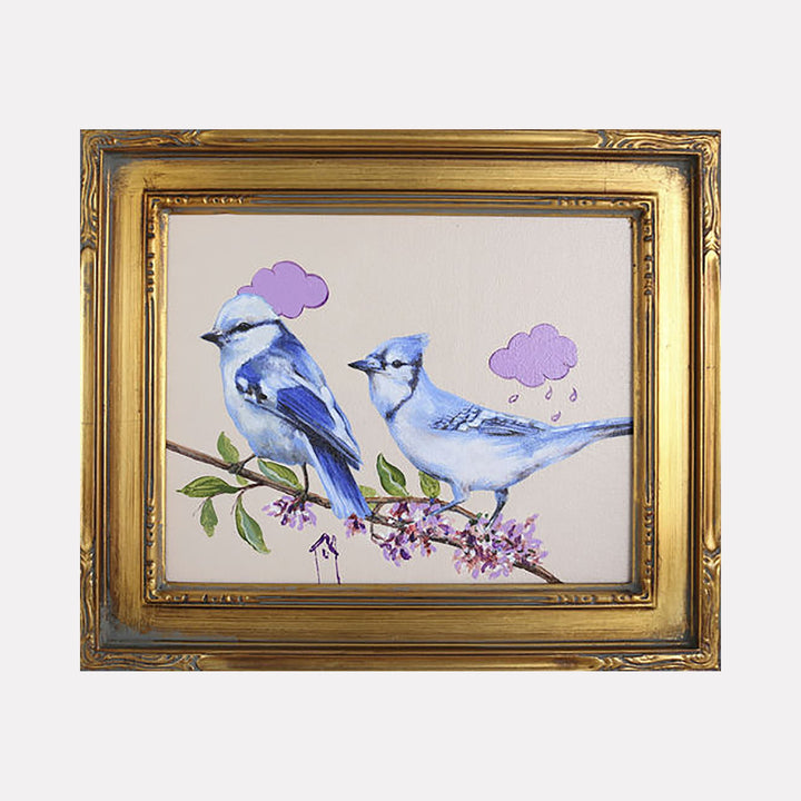 The artwork "A Home in the Woods" by Carlos Gamez de Francisco features two blue and white birds perched on a flowering branch with green leaves and purple blossoms. Above each bird are stylized purple cloud shapes, one with raindrops. The piece is framed in an ornate gold frame, showcasing soft textures and delicate brushstrokes.