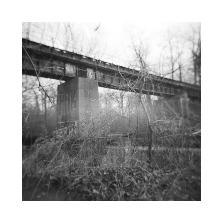 The artwork "Bikes in the River" by Kelly McGovern features a black and white photograph capturing an old bridge with large concrete pillars surrounded by leafless trees and dense brush. The image showcases a textured, moody atmosphere with soft focus, evoking a sense of quiet abandonment. Kelly McGovern.