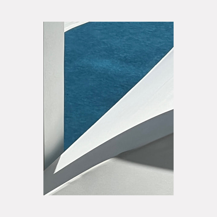 The artwork "Blanco y Azul" features an abstract composition of clean white architectural elements casting soft shadows against a vibrant textured blue background, highlighting sharp geometric shapes and smooth surfaces. The minimalist style emphasizes light and contrast. By Starr Tucker-Ortega.