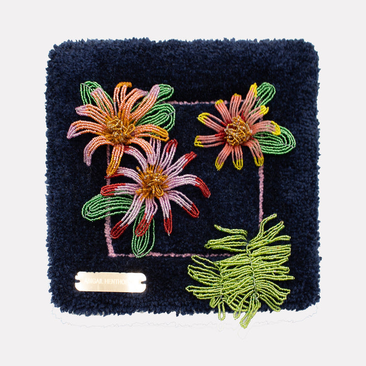 The artwork Blue Flower Arrangement by Abigail Henthorne features intricate beaded flowers in soft pinks and warm oranges with vibrant green leaves, set against a rich textured deep blue background. The detailed craftsmanship and bold contrast evoke the tactile quality and floral abstraction reminiscent of Georgia O’Keeffe’s expressive botanicals and the meticulous detail of Jan van Huysum’s still life paintings.