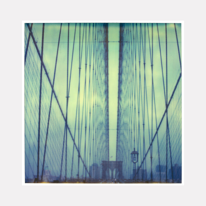 The artwork "Brooklyn Bridge (Stay)" features a soft, atmospheric view of the iconic Brooklyn Bridge with its intricate web of suspension cables framed by a misty, bluish-green palette. The texture has a vintage, almost dreamlike quality with blurred edges and subtle light reflections. By Stefanie Schneider.