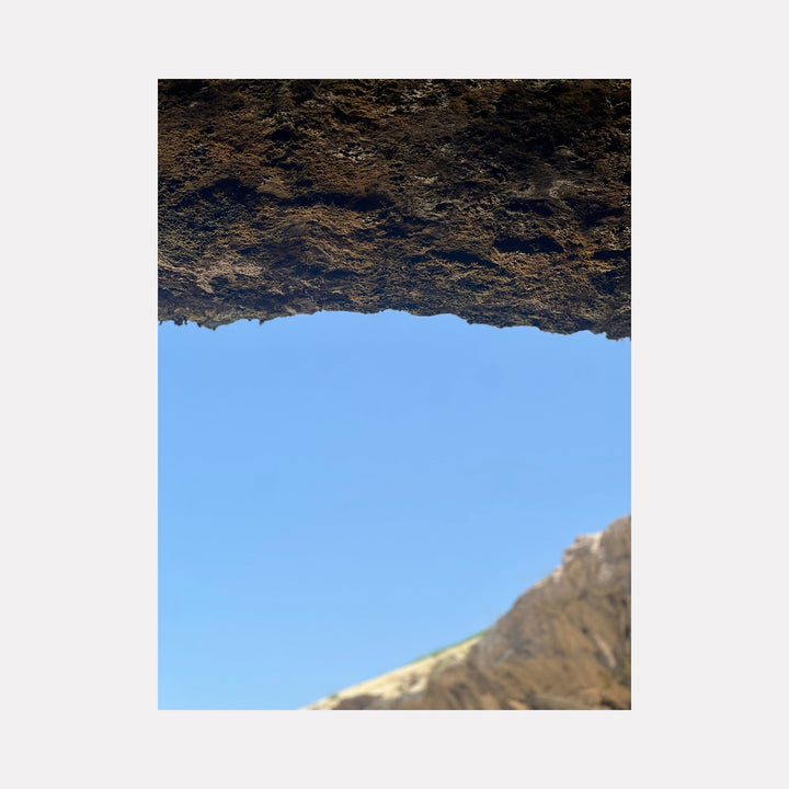 The artwork "Caven 1" by Starr Tucker-Ortega features a textured brown rocky overhang framing a clear blue sky, with a sunlit rugged mountain peak visible in the lower right corner. The contrast between the rough rock surface and smooth sky creates a natural, minimalistic composition. Starr Tucker-Ortega.
