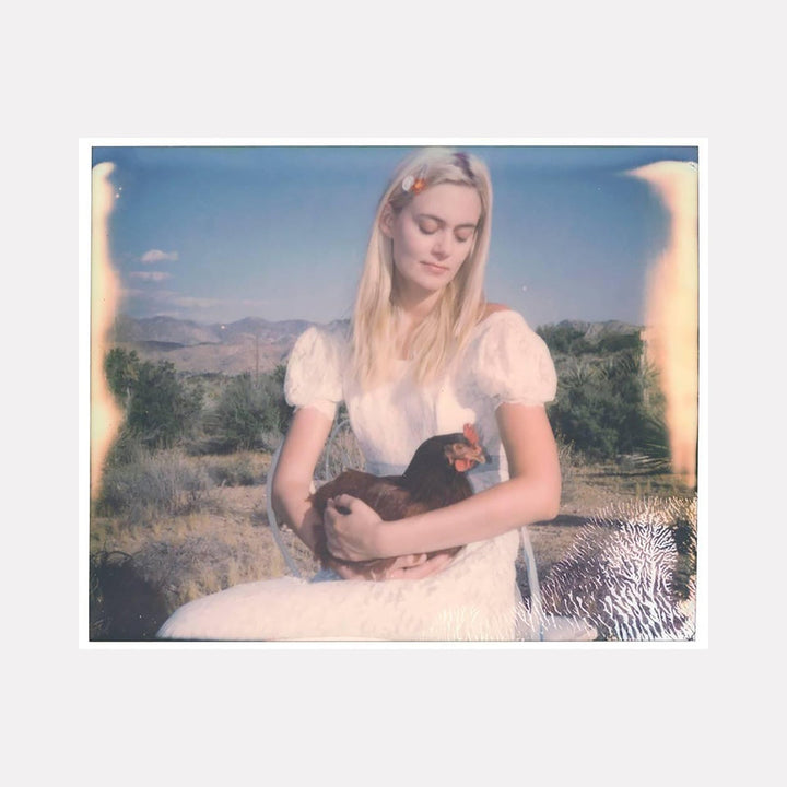 The artwork "Chicken Madonna (Chicks and Chicks and sometimes Cocks)" by Stefanie Schneider depicts a serene young woman in a white dress holding a dark rooster, set against a blurred desert landscape with muted tones. The image has a vintage, faded texture with soft lighting and subtle imperfections, creating a nostalgic and peaceful atmosphere.