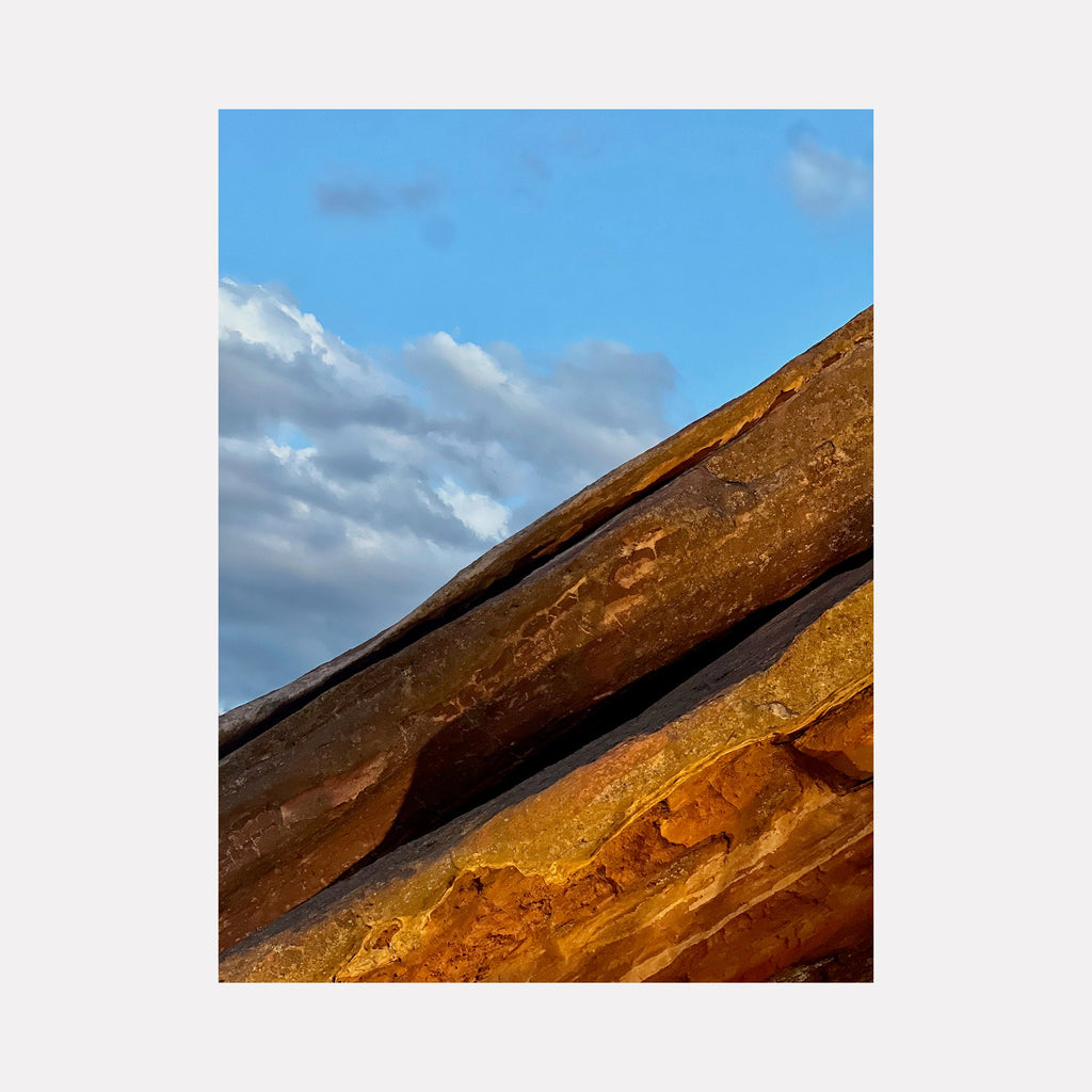 The artwork "Cretaceous 1" by Starr Tucker-Ortega depicts close-up textured rock formations with rich brown and orange hues contrasted against a bright blue sky with scattered clouds, showcasing natural geological layers and rough surfaces in a detailed, vibrant style.