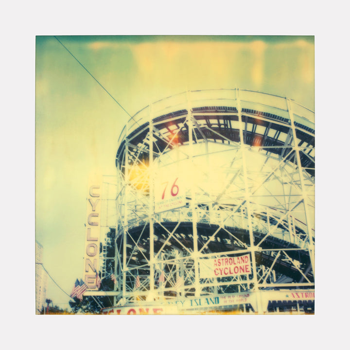 The artwork "Cyclon (Stay)" features a vintage roller coaster structure with intricate white metal framework bathed in soft, warm light tones of yellow and blue, capturing a nostalgic carnival atmosphere. The image emphasizes geometric patterns and illuminated signs, by Stefanie Schneider.