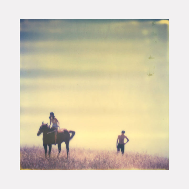 The artwork "Days of Heaven (29 Palms, CA)" by Stefanie Schneider depicts a soft, dreamy scene with muted pastel tones, featuring a person riding a horse and another standing in a field of tall grasses under a hazy sky. The image has a vintage, ethereal quality with blurred textures and subtle color gradients. Vendor: Stefanie Schneider.