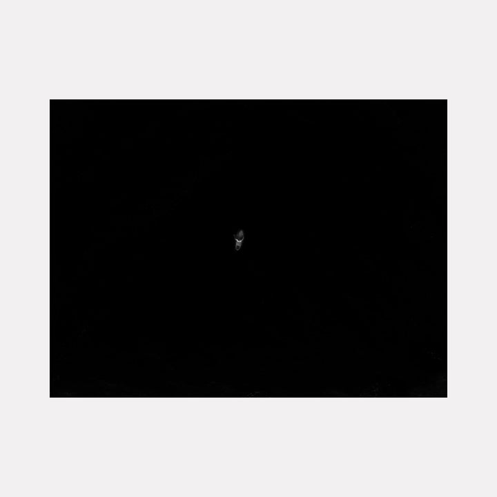 The artwork "Held by Nothing" features a minimalist composition with a solitary, faintly illuminated human face emerging from a deep black background, evoking themes of isolation and existential contemplation. The style recalls the stark contrasts and emotional depth found in works by Francis Bacon and Alberto Giacometti. Presented by Dennis Maida.