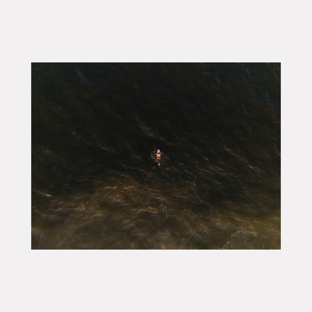 The artwork "Golden Hour Drift" depicts a lone kayaker paddling through dark, textured waters illuminated by a warm, golden light creating a serene contrast reminiscent of the atmospheric depth found in J.M.W. Turner's seascapes. The subtle interplay of light and shadow evokes a calm, meditative mood. Dennis Maida.