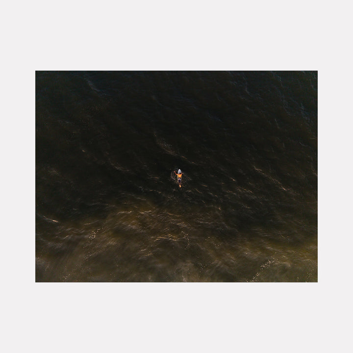 The artwork "Golden Hour Drift" depicts a lone kayaker paddling through dark, textured waters illuminated by a warm, golden light creating a serene contrast reminiscent of the atmospheric depth found in J.M.W. Turner's seascapes. The subtle interplay of light and shadow evokes a calm, meditative mood. Dennis Maida.