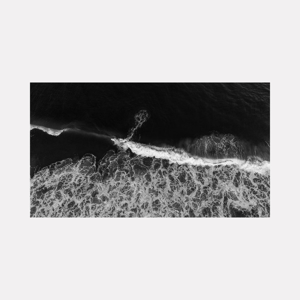The artwork "In The Break" features a monochromatic aerial view of ocean waves crashing and foaming, creating intricate swirling patterns and textures reminiscent of Japanese ink wash paintings or the dynamic fluidity found in works by Hokusai. The black and white contrast emphasizes depth and movement. By Dennis Maida.