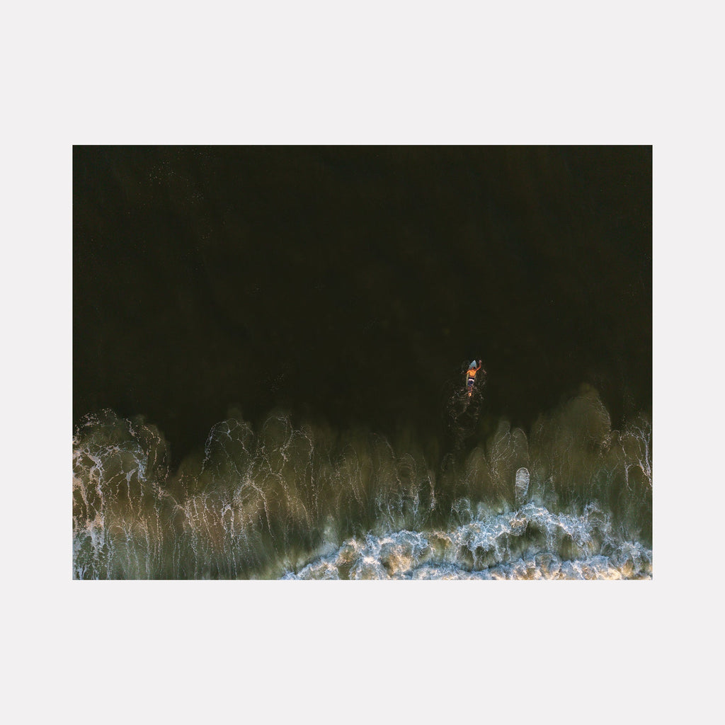 The artwork "The Push" by Dennis Maida features a lone surfer navigating dark, deep waters contrasted by foamy, white ocean waves. The image captures dynamic textures of water movement and light reflection, evoking a sense of solitude and resilience, reminiscent of the moody, naturalistic style seen in works by Hiroshi Sugimoto. Dennis Maida.