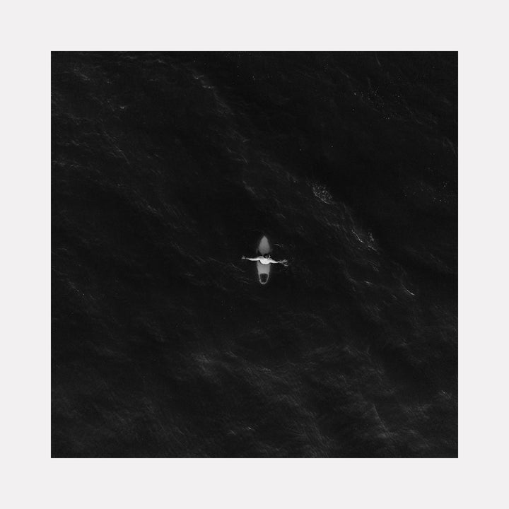 The artwork "As I Wait" features a solitary figure on a small boat amid a vast, dark, and textured ocean, rendered in monochromatic black and white tones. The high-contrast style evokes minimalism and the contemplative isolation found in works by Gerhard Richter. The subtle ripples and shadows create a mysterious, meditative atmosphere. Dennis Maida.