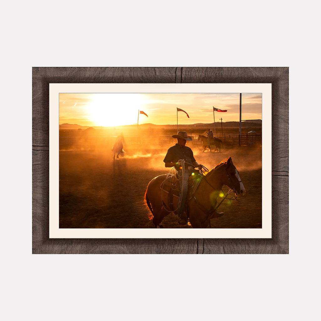 Echoes of the Frontier by Kyle Murphy portrays a warm sunset scene with cowboys on horseback riding through a dusty arena. The golden light filters through dust clouds, highlighting Western flags and evoking a nostalgic atmosphere of open plains and rugged frontier life.