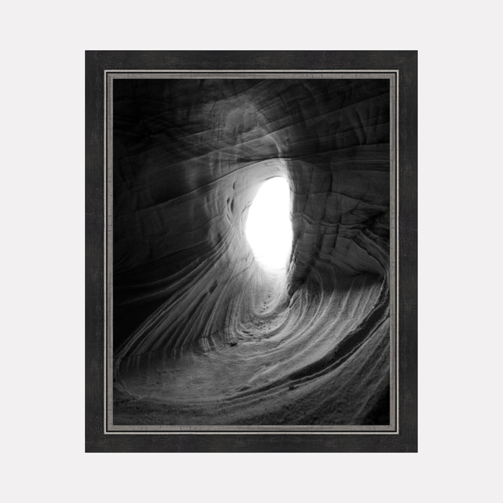 Emergence by Kyle Murphy captures a dramatic black and white view through a smooth, curved rock formation, with natural light flooding an opening that suggests a journey from darkness into illumination, framed with subtle textures that emphasize the ethereal quality of this serene landscape.