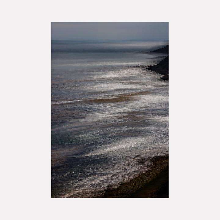 The artwork "Feral Coast" by Neil Shapiro features a moody, abstracted coastal scene with blurred, flowing textures in muted blues, grays, and earthy browns. The soft blending and atmospheric layering evoke the contemplative seascapes of Gerhard Richter, creating a serene yet dynamic visual experience grounded in nature’s raw beauty.