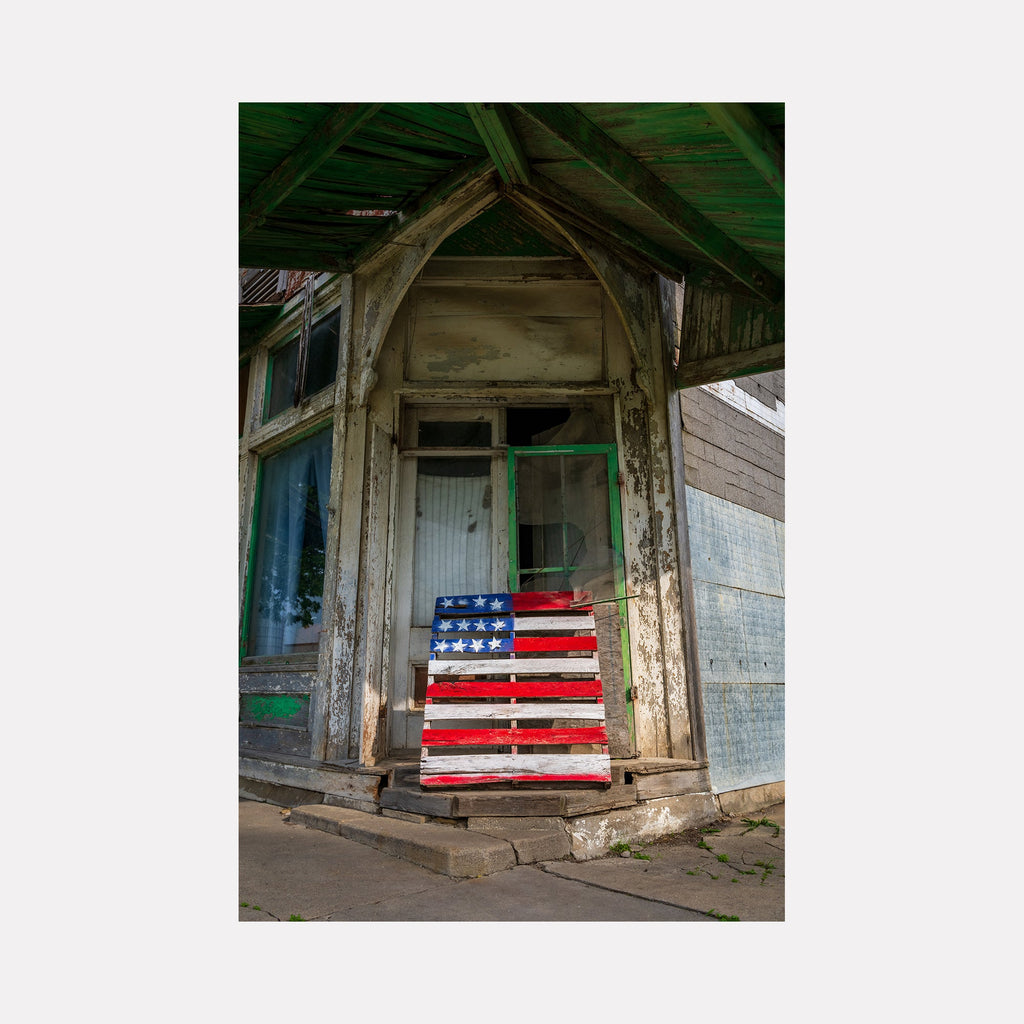 The artwork 'Faded Glory' by Dennis Maida captures a weathered wooden pallet painted as an American flag, leaning against a dilapidated storefront entrance. The composition features peeling paint, rustic architectural details, and a green-tinged wooden roof overhang. The distressed American flag palette creates a poignant contrast against the faded white clapboard siding and worn doorframe.