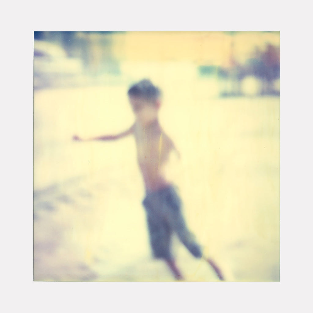 The artwork "Flying Boy (Stay)" by Stefanie Schneider depicts a blurred, soft-focus image of a young boy in motion, set against a pale, sunlit background with muted tones of yellow and blue, creating a dreamy, nostalgic atmosphere with a vintage photographic style. Stefanie Schneider.