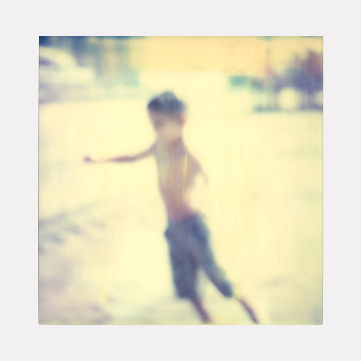 The artwork "Flying Boy (Stay)" by Stefanie Schneider depicts a blurred, soft-focus image of a young boy in motion, set against a pale, sunlit background with muted tones of yellow and blue, creating a dreamy, nostalgic atmosphere with a vintage photographic style. Stefanie Schneider.