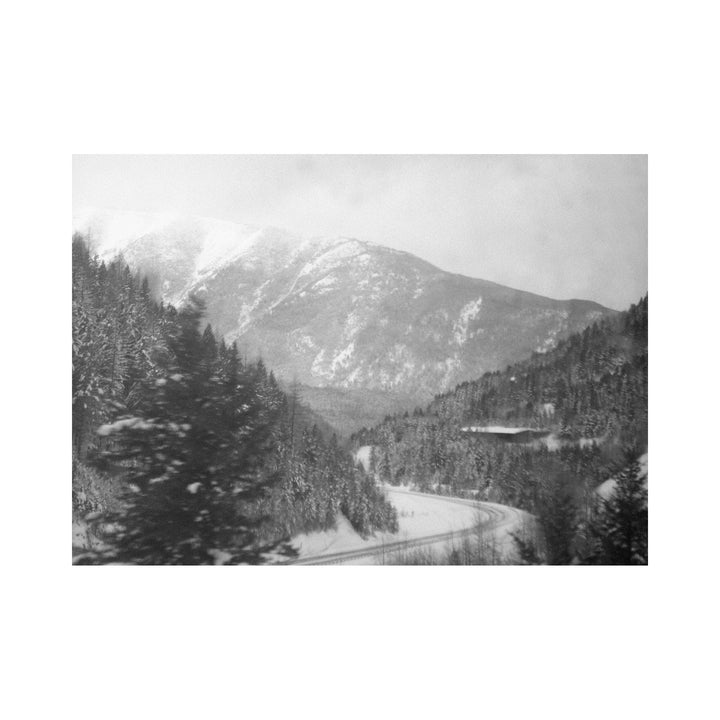 The artwork "For The Girl in The Pallet Factory" features a black and white photograph of a snowy mountainous landscape with dense pine forests and a winding road cutting through the scenery, capturing a serene and textured winter atmosphere. Created by Kelly McGovern.