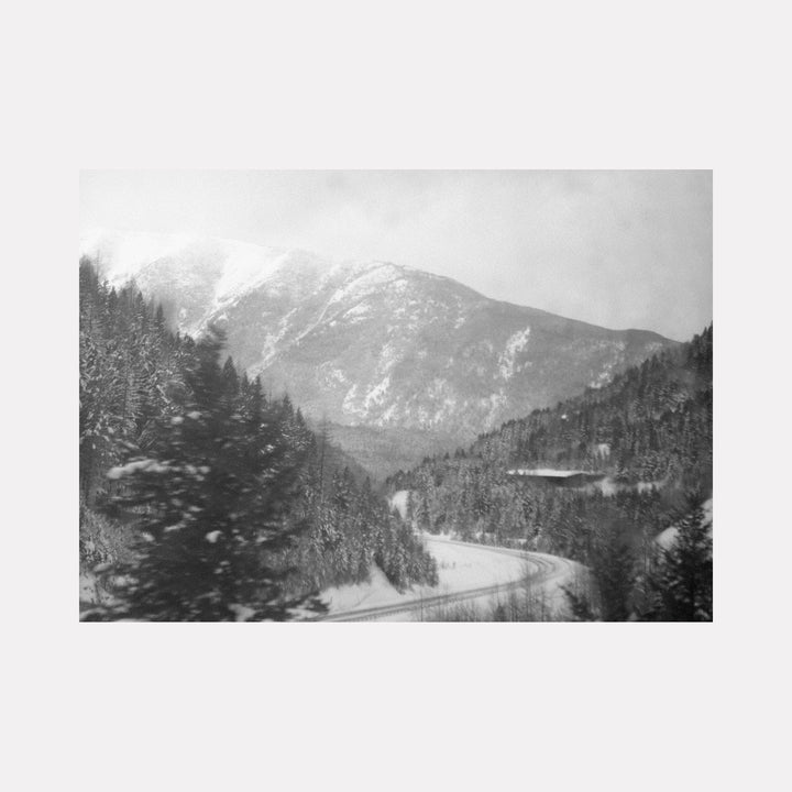 The artwork "For The Girl in The Pallet Factory" by Kelly McGovern features a monochromatic winter landscape with snow-covered pine trees and a winding road leading toward misty, rugged mountains. The soft, blurred textures evoke a dreamy atmosphere reminiscent of early 20th-century photographers like Ansel Adams, blending realism with poetic ambiance in a timeless natural scene.