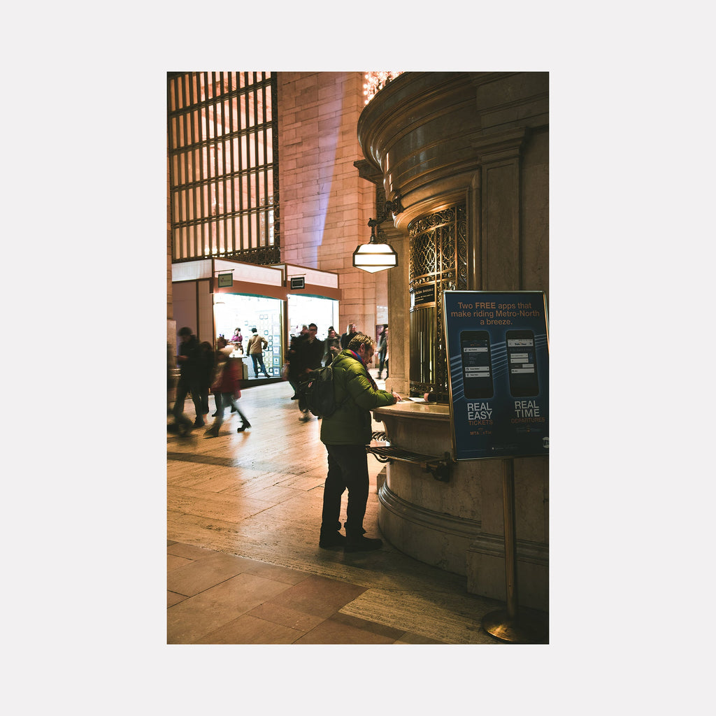 The artwork 'Get Me Home' by Dennis Maida captures a moody scene inside Grand Central Terminal, featuring warm-lit architectural elements, marble columns, and an ornate information booth. The photograph showcases classic Beaux-Arts interior design with golden lighting, arched windows, and stone flooring, creating a nostalgic urban atmosphere. A figure stands at the booth while commuters move through the background. By Dennis Maida.
